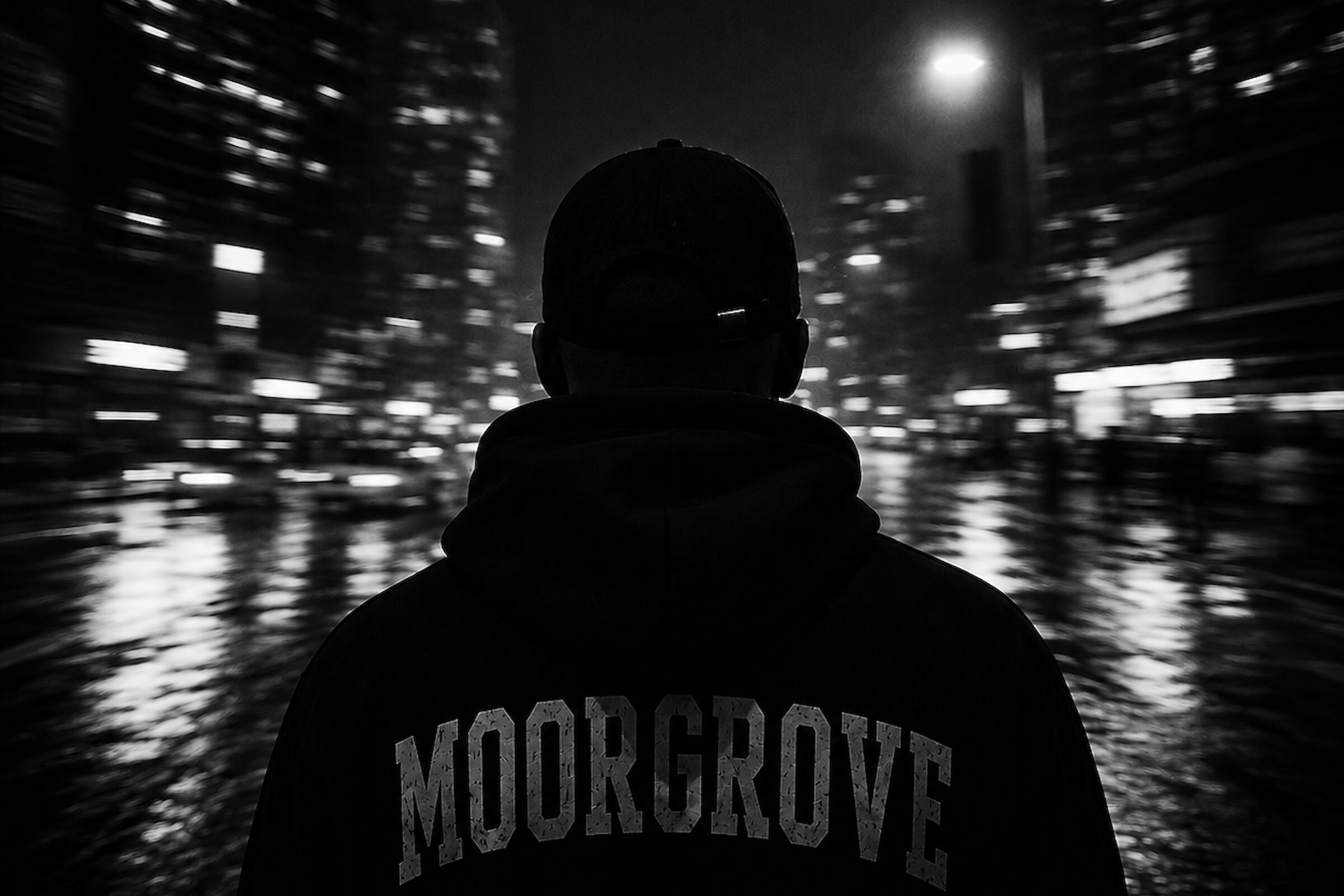 Figure in a MOORGROVE hoodie facing a rain-lit city at night
